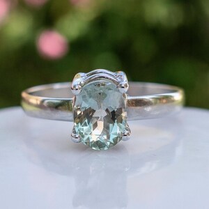 May include: A silver ring featuring an oval, pale green gemstone. The gemstone is set with four prongs. The ring is displayed on a white surface with a blurred green and pink background.
