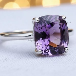 May include: A silver ring featuring a large, cushion-cut, purple amethyst gemstone. The gemstone is set with four prongs, and the band is smooth and polished. The ring is displayed on a white surface.