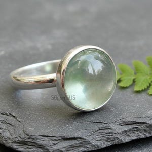 May include: A silver ring featuring a large, round, pale green gemstone. The gemstone is set in a simple bezel setting, and the band is smooth and polished. The ring is displayed on a dark, textured surface.