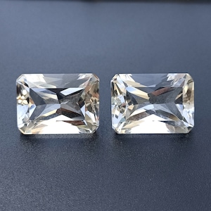 Puede incluir: Dos gemas transparentes, rectangulares y facetadas con un ligero tono amarillo. Las gemas están colocadas sobre un fondo gris oscuro.