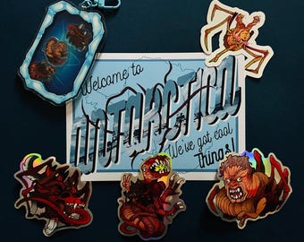 May include: A set of stickers featuring various creatures, including a dragon, a monster, and a crab. The stickers are holographic and have a blue background. The stickers are arranged on a blue surface with a postcard that says "Welcome to  " in a retro font.