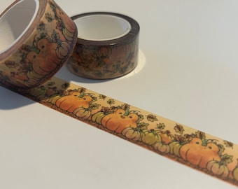 May include: Two rolls of washi tape with a fall theme. The tape features a pattern of brown and orange pumpkins with green leaves. The tape is on a white surface.