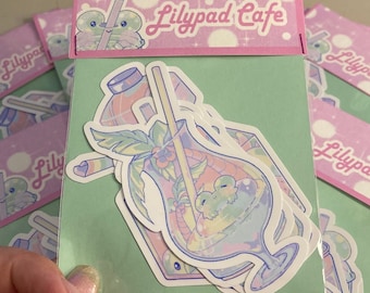 May include: A stack of stickers featuring a cartoon frog in a glass of tropical fruit punch. The stickers are printed on white paper with a light green background and have a pink border with the text "Lilypad Cafe".