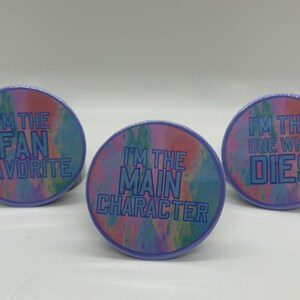 May include: Three round buttons with a colorful background and blue text. The text on the buttons reads "I'm the Fan Favorite", "I'm the Main Character", and "I'm the One Who Dies".