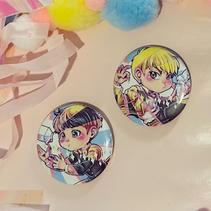 May include: Two circular badges featuring anime-style illustrations of a male character with blonde and black hair. The badges have a glossy finish and are set against a light background with pastel decorations.