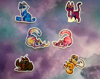 May include: A collection of six cat stickers with a celestial theme. Each sticker features a cat in various poses, adorned with crowns and accessories. The cats are depicted in shades of blue, purple, pink, and gold, set against a galaxy-themed background.