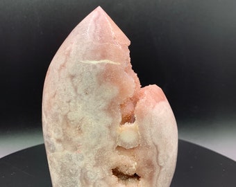May include: A large, pink, crystal geode with a pointed top and a large opening revealing the inner crystals. The geode has a white and pink marbled appearance.
