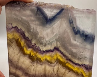 May include: A slab of fluorite with a layered pattern of purple, yellow, and blue. The fluorite has a smooth, polished surface.
