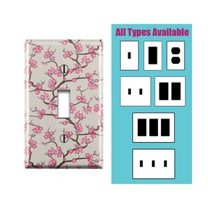 Peut inclure: Une plaque de prise de courant avec un motif de fleurs de cerisier roses sur un fond beige. La plaque est présentée dans une configuration à un bouton, mais le texte "Tous les types disponibles" indique que d'autres configurations de plaques de prise de courant sont disponibles.