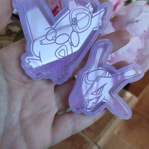Op de afbeelding: Twee transparante paarse acrylcharms met lijntekeningen van Pokémon-personages. De eerste charm is van een Pokémon met een groot hoofd en een klein lichaam, die een kleinere Pokémon in zijn armen vasthoudt. De tweede charm is van een Pokémon met een groot hoofd en een puntige snuit.