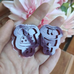 Puede incluir: Dos moldes de silicona transparentes para fundición de resina, con un diseño de niña y niño. Los moldes están etiquetados con "MOONLIGHTMOLDS".