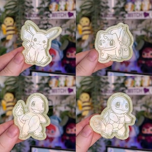 Könnte beinhalten: Ein Set aus vier irisierenden, halbtransparenten Harzanhängern mit eingravierten Umrissen der Charaktere Pikachu, Bisasam, Glumanda und Schiggy. Jeder Anhänger hat eine glatte, glänzende Oberfläche mit einem perlmuttartigen Schimmer.