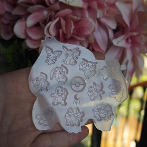 May include: A clear silicone mould with various Pokémon character designs. The mould includes images of Pikachu, Charmander, and others, along with a Poké Ball symbol. The mould is held in a hand, with pink flowers in the background.