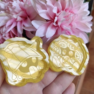 Puede incluir: Dos piezas de resina doradas, estilo caricatura, con veteado blanco. Una presenta un personaje con sombrero y lengua fuera, la otra un personaje con un diseño complejo. Flores rosas en el fondo.