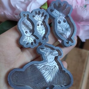 Puede incluir: Tres moldes de resina para manualidades. Los moldes son transparentes y muestran el diseño de un pájaro y dos pequeñas criaturas con ojos grandes. Los moldes están en una mano.