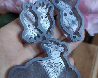 May include: Three resin molds for crafting. The molds are clear and show the design of a bird and two small creatures with large eyes. The molds are on a hand.