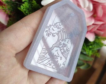 Puede incluir: Un llavero de resina transparente de forma rectangular con un contorno blanco de la Gran Ola de Kanagawa, una famosa estampa japonesa en madera.