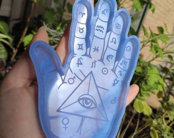 May include: A light blue resin hand tray with an all-seeing eye and astrological symbols. The tray is designed to hold small items like jewelry or keys.