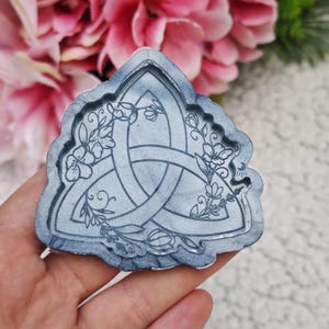 Puede incluir: Un molde de resina gris de un nudo triquetra con diseños florales. El molde tiene forma de triángulo.