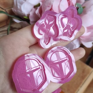 Pode incluir: Dois moldes de silicone rosa para fundição de resina. Um molde apresenta um design de capacete mandaloriano, o outro apresenta um design do símbolo mandaloriano. Ambos os moldes têm o texto "Moonlightmolds" impresso neles.