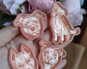 May include: Four peach-colored dog-shaped resin charms. Each charm features a different detailed illustration of a dog's face or body. The charms are held in a hand, with a blurred floral background.