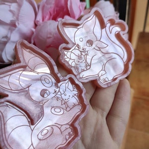 Può includere: Due ciondoli in resina rosa con un'illustrazione di cartone animato di un Eevee rosa chiaro che tiene un Eevee rosa scuro più piccolo. I ciondoli sono contornati da un rosa più scuro.