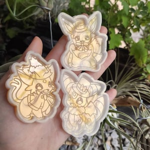 May include: Three clear resin molds of Pokemon characters wearing hats. The molds are translucent and have a gold shimmer. The characters are Eevee, Vaporeon, and Flareon.