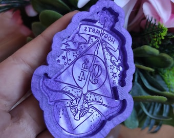 May include: A purple silicone mold featuring the Deathly Hallows symbol, a wand, and the word "HOGWARTS" in reverse. The mold has a decorative border with stars and banners. The design is for crafting or resin art.