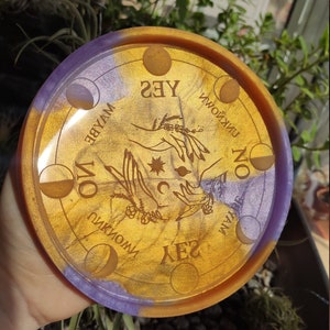 May include: A round, gold and purple resin tray with a moon phase design and the words "YES", "MAYBE", "NO", "UNKNOWN", and "UNKNOWN" around the edge.