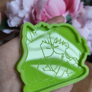 May include: Green silicone mold with a raised design of two cartoon characters kissing. The characters are outlined in white.