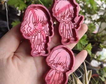 May include: Three resin character charms in a hand. The charms are red and feature cartoon-style characters with black outlines. The characters are a girl with long curly hair, a boy with short hair and glasses, and a boy with short hair.
