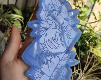 Puede incluir: Un molde de resina azul claro con un diseño floral con una luna, cristales y sol. El molde se sostiene en una mano, con los detalles del diseño en un tono más claro de azul. El molde es para manualidades.