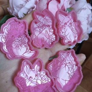 Può includere: Cinque ciondoli in resina rosa con diversi personaggi dei cartoni animati con accenti floreali. I ciondoli sono in una mano, e lo sfondo è un fiore rosa.