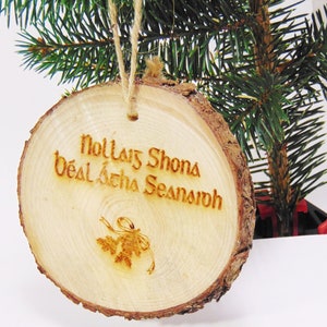 Pode incluir: Um ornamento redondo de madeira com uma borda de casca marrom rústica. O ornamento é decorado com um design gravado a laser de um sino e o texto "Nollaig Shona Déas Seachta Seansr  íoch".