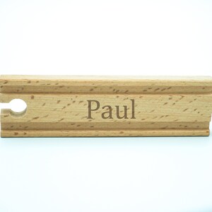 Op de afbeelding: Een houten treinbaanstuk met de naam "Paul" gegraveerd in een lichtbruin lettertype. De baan heeft een lichte houtnerf en een sleuf aan één uiteinde en een pen aan de andere kant. De achtergrond is wit.