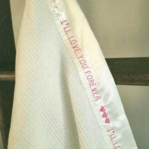 Op de afbeelding: Een witte deken met wafelstructuur en een gladde satijnen rand. De satijnen rand is geborduurd met de roze tekst I'LL LOVE YOU FOREVER, gevolgd door drie kleine roze hartjes, wat een decoratief detail vormt langs de stoffen rand.