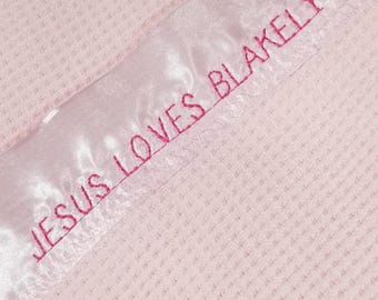 May include: A light pink waffle-textured blanket featuring a smooth satin border. The satin trim is embroidered with the text JESUS LOVES BLAKELY in dark pink thread. The fabric has a consistent grid-like pattern across the main surface area.