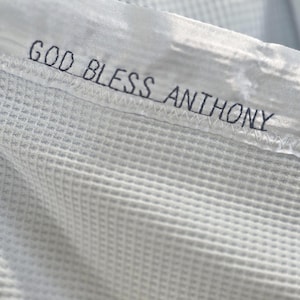 May include: A white waffle-textured fabric featuring dark blue embroidered text that reads GOD BLESS ANTHONY. The embroidery includes two small heart icons on the left side and a small car icon on the right side, finished with a zigzag stitch.