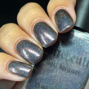 May include: Close-up of manicured nails painted with a dark, shimmering nail polish. The polish has a holographic effect, reflecting multiple colors. The bottle of nail lacquer is partially visible, with the text "Cult" and "nail lacquer".