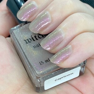 Puede incluir: Una botella de esmalte de u&ntilde;as de vidrio transparente con una etiqueta plateada que dice "Witchy Nails" y "Capricornio". La botella contiene un esmalte de u&ntilde;as gris claro con un acabado brillante. La botella es de 15 ml.