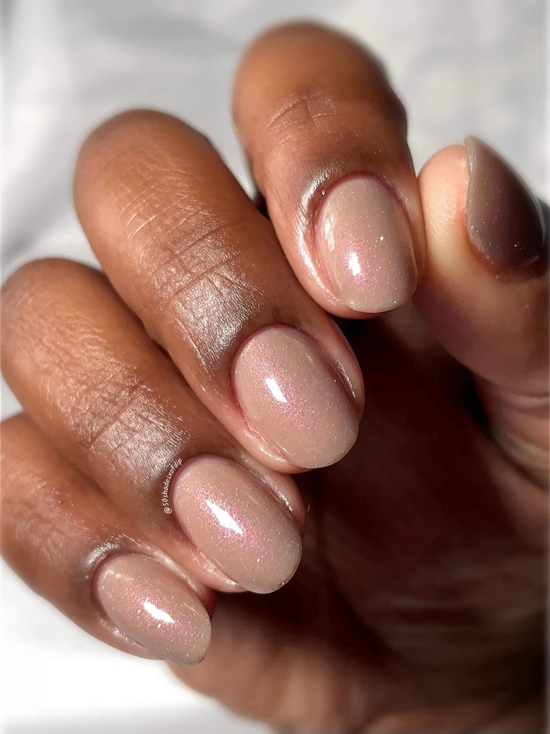 Puede incluir: Primer plano de una mano con esmalte de u&ntilde;as de color nude con un brillo sutil. Las u&ntilde;as son de forma ovalada y tienen un acabado brillante.