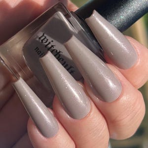 May include: A glass bottle of Witchcult nail polish with a black cap, held alongside long, coffin-shaped fingernails painted in a matching shimmery taupe-grey color. The bottle label features the text Witchcult in a gothic-style font.