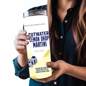 Könnte beinhalten: Eine weiße Cutwater Lemon Drop Martini-Dose mit silbernem Deckel wird gehalten. Die Dose zeigt den Text "CUTWATER LEMON DROP MARTINI" in Blau, mit einer Zitrone und einer Eiswürfel-Grafik. Die Dose hat 11 % Alkohol.