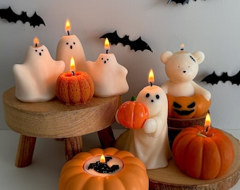 May include: A collection of Halloween-themed candles. The candles are shaped like ghosts, pumpkins, and a bear, with lit flames. The color palette includes white, orange, and black, with decorative bat cutouts in the background. The candles are arranged on a wooden surface.