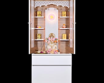 May include: A white and wood-toned cabinet with open doors, featuring shelves holding religious items. A central deity statue is surrounded by flowers. The top of the cabinet has text in a foreign language.