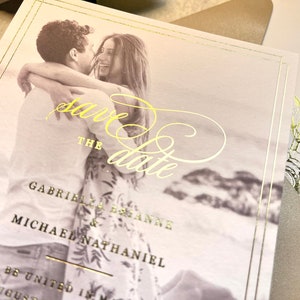 May include: Save the date card with a gold foil design. The card features a photo of a couple embracing, with the text "Save the Date" in gold foil script. The couple's names are listed below the photo: Gabrielle & Michael Nathaniel.