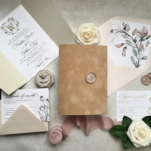 May include: Wedding invitation suite with a cream and blush color scheme. The invitation features a gold crest with the initials "W" and "V". The RSVP card features a floral design with a black and white illustration. The outer envelope is a light brown color with a wax seal.