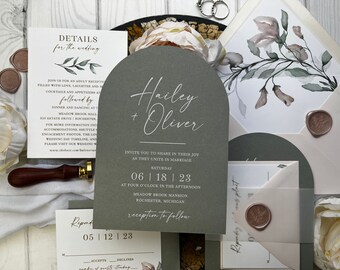 May include: A wedding invitation suite with a sage green arch-shaped card with white calligraphy that reads "Hailey + Oliver". The invitation details the wedding date, time, and location. The invitation is accompanied by a white envelope with a floral design and a wax seal.