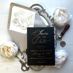 May include: A black "Save the Date" card with gold lettering, featuring the names Crystal & Brandon and the wedding date. An envelope with gold rose illustrations, scissors, and white flowers are also present.