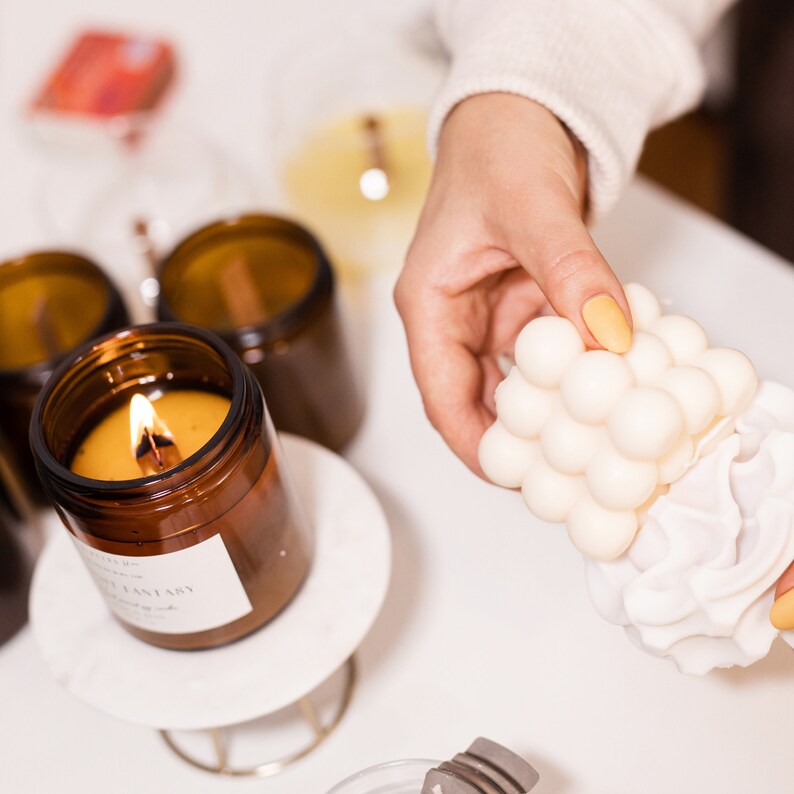 Proceso de fabricación de una vela: manos femeninas retiran una vela moldeada, al lado una jarra de ámbar con una vela encendida.